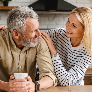 Woman touching man’s shoulders as they smile at each other at kitchen counter