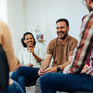 4 adults sitting on chairs laughing and talking in a white room