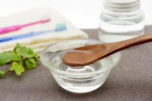 A wooden spoon over a small jar of melted coconut oil sitting on a brown tablecloth. Toothbrushes lay blurred in the background.