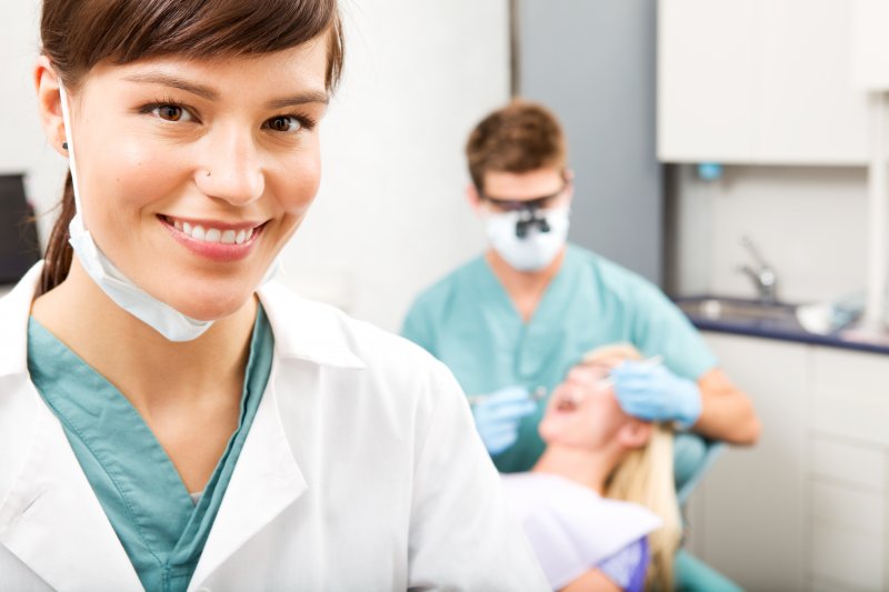 a dental assistant smiling while the dentist examines a female patient’s smile in the background